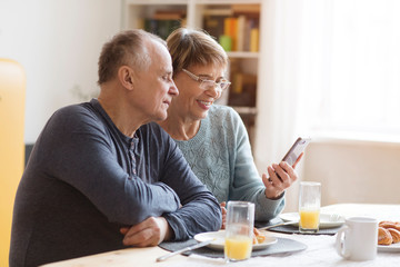 Senior couple eating breakfast and using smartphone at home