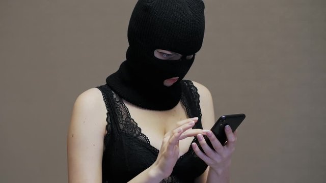 Wife or girlfriend in black mask balaclava use mobile phone of her husband, woman with big breasts dressed in sexual underwear, surprised by treason