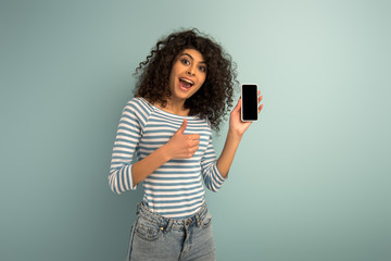 happy mixed race girl showing thumb up while holding smartphone with blank screen on grey background