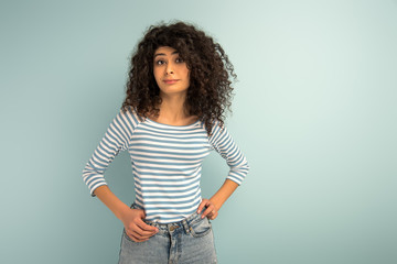 skeptical mixed girl looking at camera while holding hands on hips on grey background