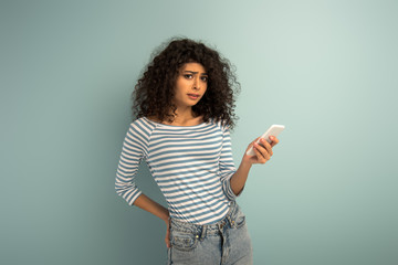 displeased mixed race girl looking at camera while using smartphone on grey background
