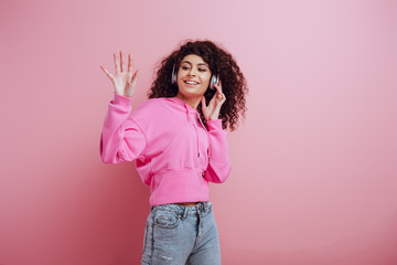 cheerful bi-racial girl waving hand while listening music in wireless headphones on pink background