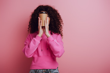 mixed race girl in pink hoodie obscuring face with paper cup on pink background
