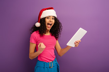 cheerful mixed race girl in santa hat showing digital tablet with blank screen on purple background