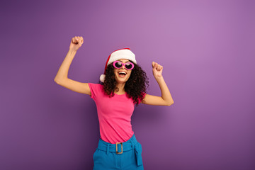 Fototapeta na wymiar cheerful bi-racial girl in santa hat and sunglasses dancing on purple background