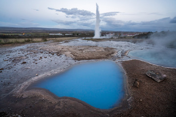 The famous geysir/Strokur with high burst, in Haukadalur valley near Reykjavik in Iceland during blue hour with no people around. Clear blue hot pool used as foreground. Tourist attraction concept.