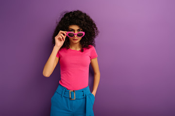 confident bi-racial girl touching sunglasses while looking at camera on purple background
