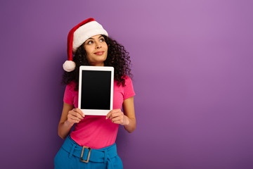 dreamy mixed race girl in santa hat looking away while showing digital tablet with blank screen on...