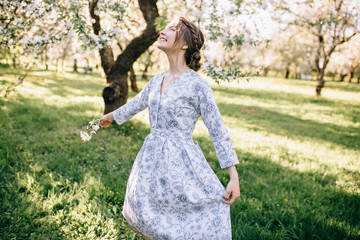 Obraz premium Portrait of young beautiful girl in white lace dress in apple garden, in the tree crown, fingers near mouth