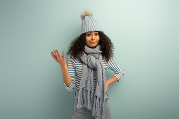 offended bi-racial girl in warm hat and scarf looking at camera on grey background