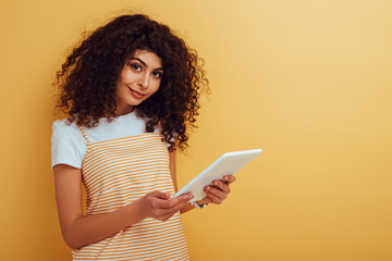 pretty, smiling mixed race girl looking at camera while holding digital tablet on yellow background