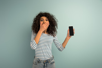 shocked bi-racial girl covering mouth with hand while showing smartphone with blank screen on grey background
