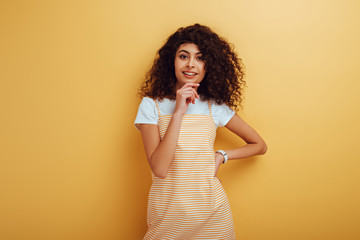 smiling bi-racial girl looking at camera while standing with hand on hip on yellow background