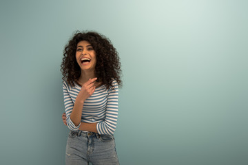excited bi-racial girl laughing while pointing with finger on grey background - obrazy, fototapety, plakaty