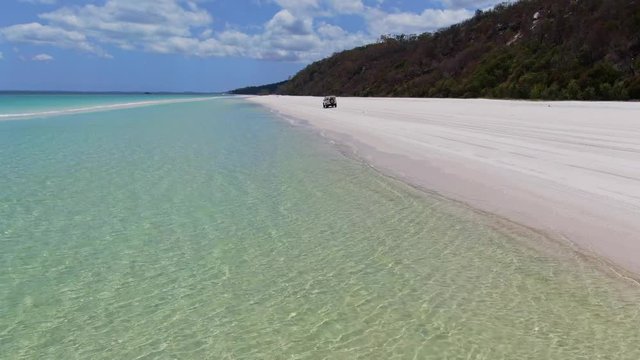 Stunning aerial drone footage of a white coloured four-wheel drive off-road vehicle driving along the vast beach on the West Coast of Fraser Island, Queensland, Australia.Clear water and gentle waves.