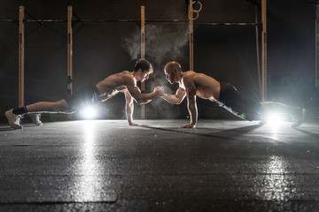 Athletic sportsmen doing push ups together and holding hands in crossfit gym - obrazy, fototapety, plakaty