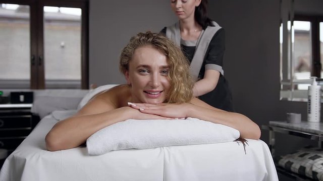 Front view of smiling charming blond woman which relaxing during body massage in beauty center