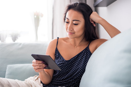 Young Asian Woman Sitting On Sofa Using Reader Tablet And Reading A Ebook At Home