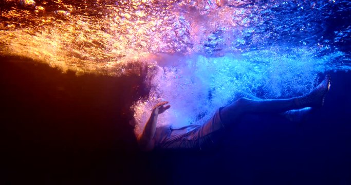 darkness with red and blue rays of light. a woman in silver clothing falls under the water in a stream of bubbles. waves are on the surface