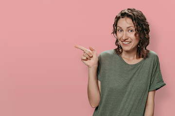 Positive smiling woman indicates with index finger at blank copy space, has amazed happy expression. Girl shows something exciting, isolated on pink background. Space for advertisement or promotional