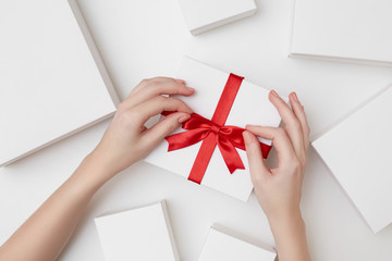 A white present box with red ribbon and women's hands tying a bow surrounded with white boxes on...