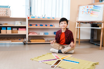 Smiling asian kid looking at camera by educational game on floor in montessori school