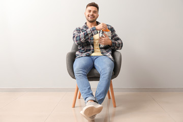 Handsome young man with tasty potato chips sitting on chair near light wall