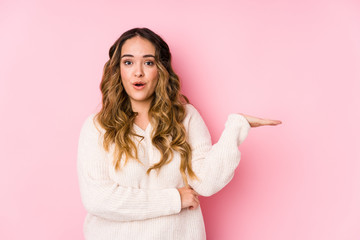Young curvy woman posing in a pink background isolated impressed holding copy space on palm.