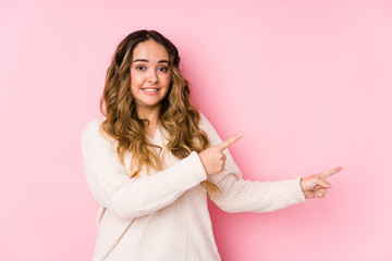 Young curvy woman posing in a pink background isolated shocked pointing with index fingers to a copy space.