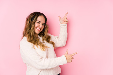 Young curvy woman posing in a pink background isolated pointing with forefingers to a copy space,...