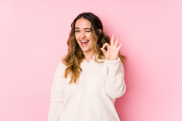 Young curvy woman posing in a pink background isolated winks an eye and holds an okay gesture with hand.