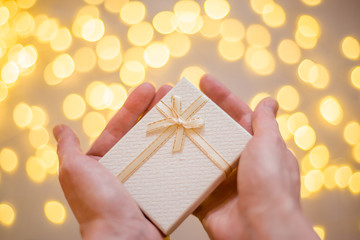 Beige box with a gold ribbon and a bow in hand on the background of bokeh lights.