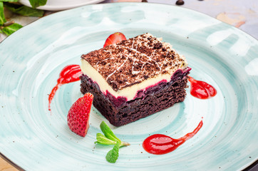 Delicious piece of chocolate cake with sour cream, cherry impregnation, mint, strawberry on a blue plate, close-up on a background. Homemade dessert with cream and cherry