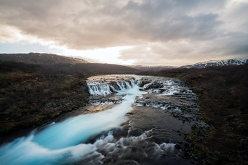 Fototapeta na wymiar Bruarfoss waterfall in Icelandic scenery during sunset and cloudy sky. Turquoise cascade and golden warm light with snow covered mountains in the background.