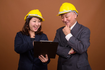 Mature Asian businessman and mature Asian businesswoman with hardhat reading on clipboard together