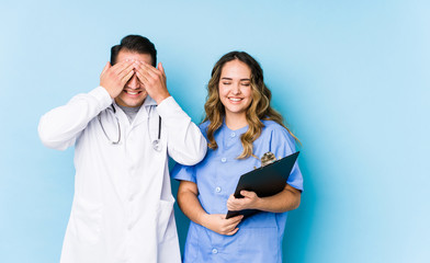 Young doctor couple posing in a blue background isolated covers eyes with hands, smiles broadly waiting for a surprise.