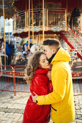 young romantic couple in love wearing in bright yellow and red down jackets hugging on french carousel background.