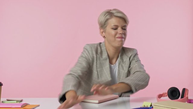 Nice young woman turns the pages of a book then throws everything off the table and start crying isolated over pink background