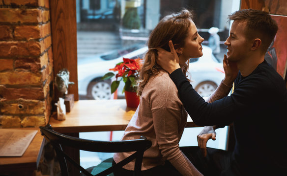 young man and woman tenderly hugges and touches each other in cozy cafe in front of big window.