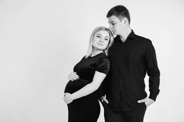 Pregnant woman and her husband hugging and holding onto your stomach in black clothes on a white background. Black and white picture. Beautiful married couple. Copy, empty space for text