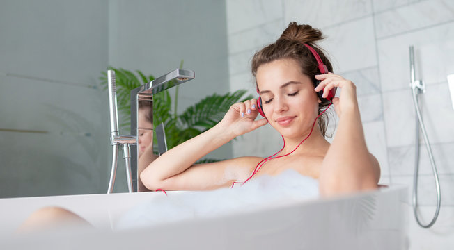 Girl with headphones listening to music while taking bath with soap foam bubbles at home bathroom.