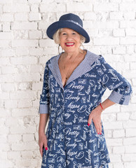 Fashionable elderly woman in a blue dress and hat against a white brick wall background smiling and posing