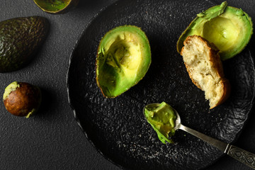 Avocado halves, bread, spoon on black plate on dark background, top view