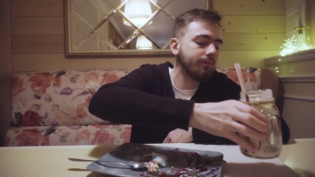 A bearded guy drinks a milkshake and eats cotton candy at a restaurant.