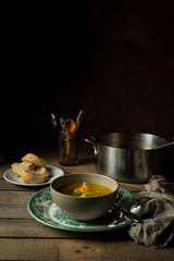 Still life with homemade chicken pasta soup, cooking pan, bread, cutlery in a glass on wooden rustic background