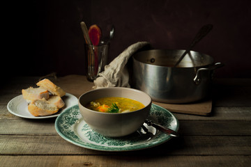 Still life with homemade chicken pasta soup, bread, metal pan, cutlery in a glass on wooden rustic background