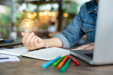 A business women hand holding a light bulb to save energy concept at home office, with computer laptop and blank notebook on workplace. A growth of thinking for creative ideas