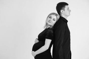 Pregnant woman and her husband stand with your back to each other in black clothes on a white background. Black and white picture. Beautiful married couple. Copy, empty space for text
