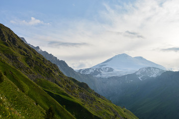 huge snow mountain Elbrus in the distance at sunset with greenery and small trees in the foreground and a glacier with waterfalls of clean drinking water