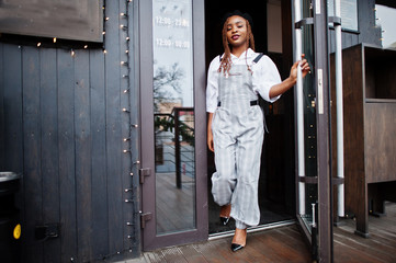 African american woman in overalls and beret walking out the door in outdoor terrace with christmas decorations.